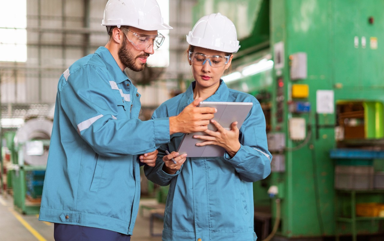Two industrial engineers in blue uniforms and white safety helmets reviewing information on a digital tablet in a manufacturing plant. They are wearing safety glasses and standing in front of machinery, discussing work-related tasks