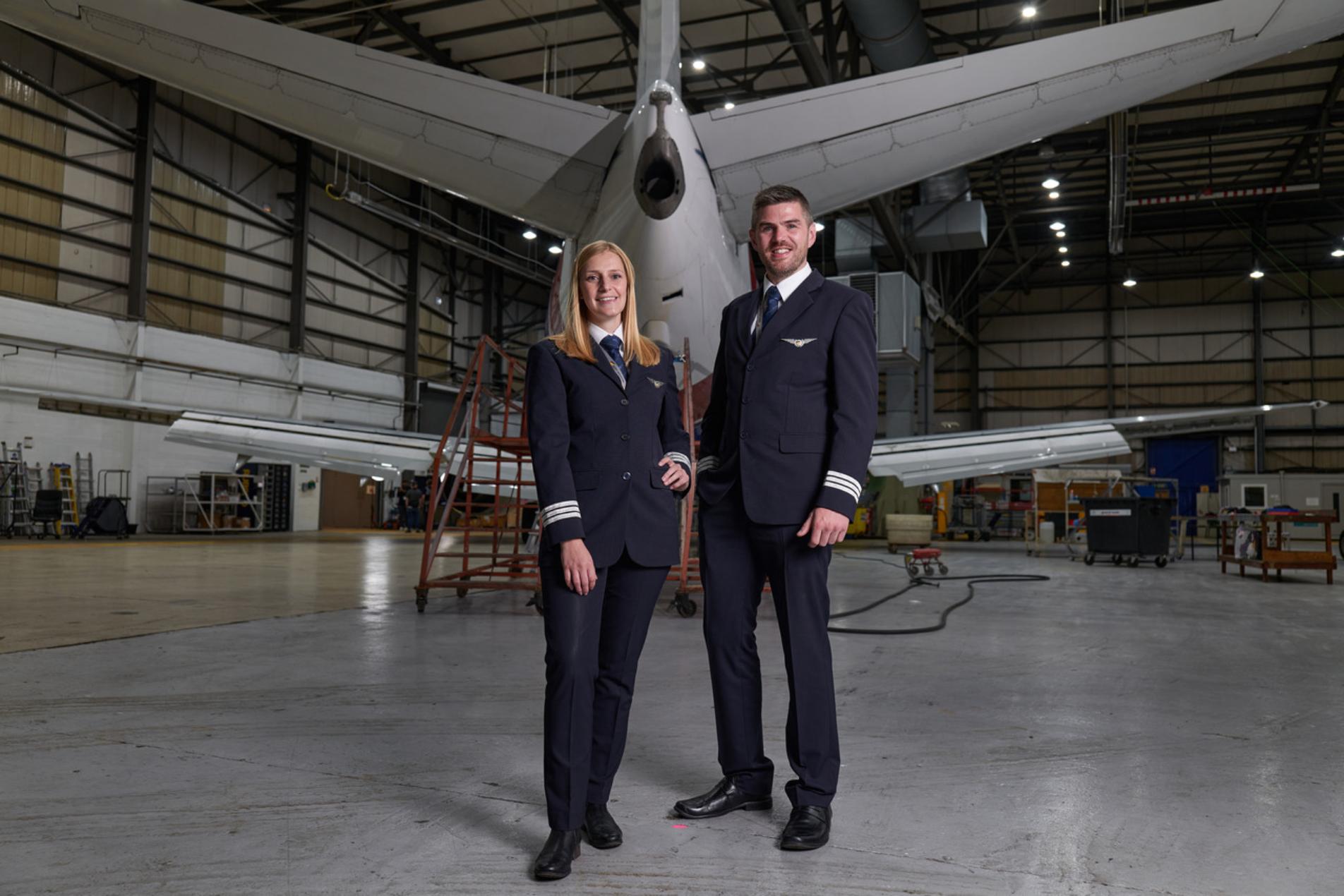 Two West Atlantic pilots in full uniform standing confidently inside an aircraft hangar. A large airplane is in the background, surrounded by maintenance equipment and scaffolding, indicating a setting of aircraft maintenance or inspection