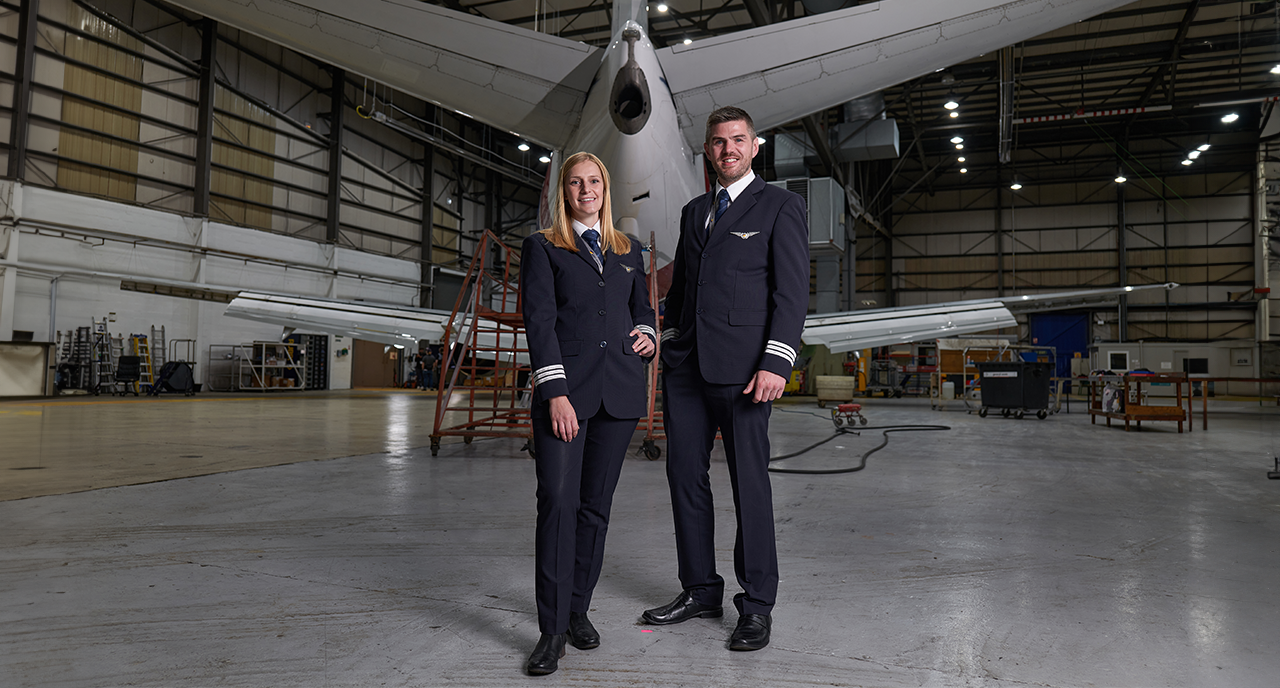 Two pilots in full uniform standing confidently inside an aircraft hangar. A large airplane is in the background, surrounded by maintenance equipment and scaffolding, indicating a setting of aircraft maintenance or inspection.