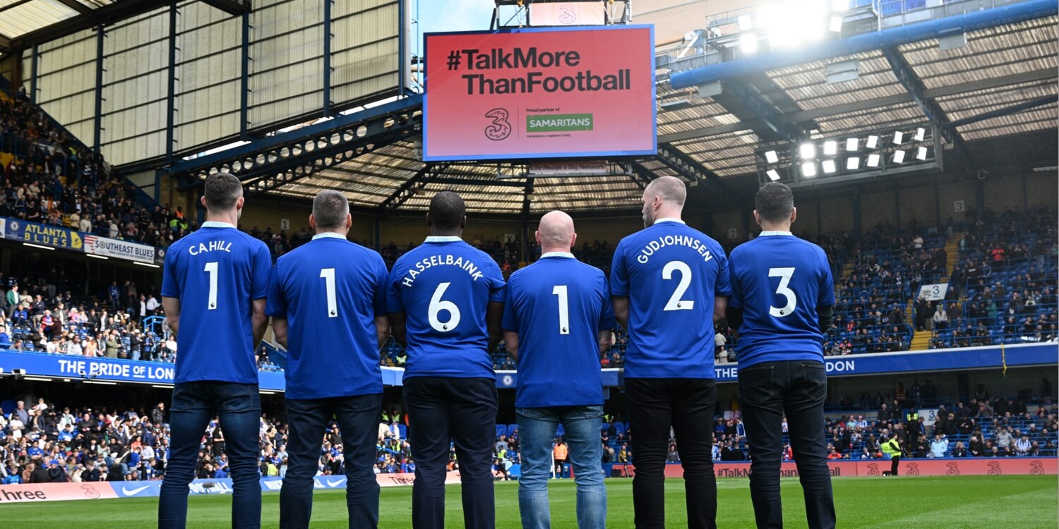 6 men with the Samaritans number 116123 on their shirts facing the talk more than football banner at Chelsea FC home ground.